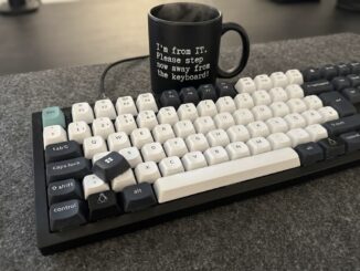 Mechanische Tastatur mit schwarz-weißen Tastenkappen auf einer grauen Filzunterlage. Unten links sitzt eine dunkle Taste mit Tux-Logo anstelle der Windows-Taste. Eine abgezogene schwarze Kappe mit Windows-Logo liegt lose auf den Tasten. Dahinter steht eine schwarze Tasse mit der Aufschrift: "I'm from IT. Please step now away from the keyboard!"