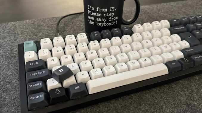 Mechanische Tastatur mit schwarz-weißen Tastenkappen auf einer grauen Filzunterlage. Unten links sitzt eine dunkle Taste mit Tux-Logo anstelle der Windows-Taste. Eine abgezogene schwarze Kappe mit Windows-Logo liegt lose auf den Tasten. Dahinter steht eine schwarze Tasse mit der Aufschrift: "I'm from IT. Please step now away from the keyboard!"